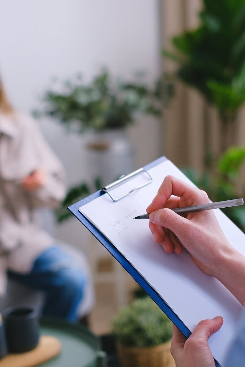 Unrecognizable professional female psychologist writing on clipboard while sitting against client on blurred background during psychotherapy session in light office