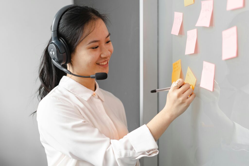 pexels photo 7682468 7682468 Young female call center agent wearing a headset, writing on sticky notes indoors.